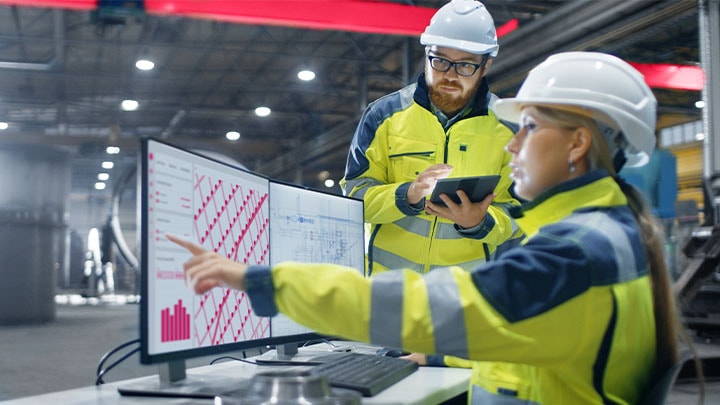 Two workers in high visibility jackets, in a warehouse, talking to each other infront of a computer