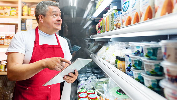 A supermarket worker with a red apron, looking at a shelf of food, holding a tablet