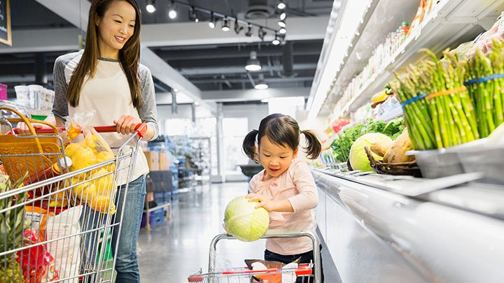 A woman smiles as her daughter places a cabbage in her own shopping cart