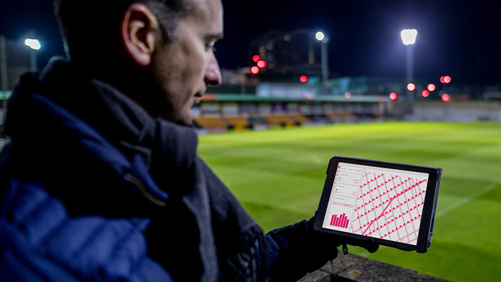 A football club manager, standing next to a pitch, looking at a tablet