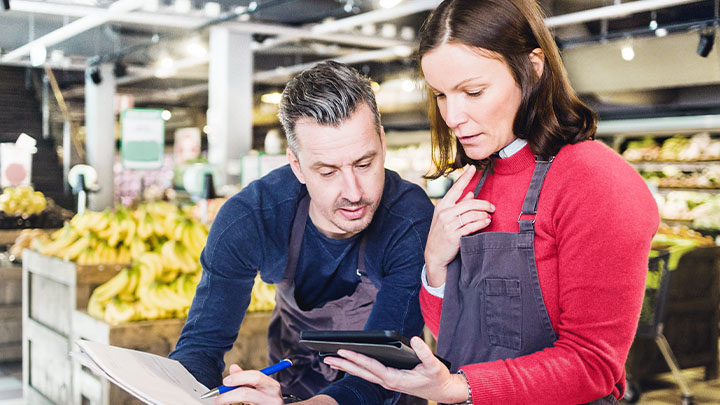 Twee collega's in een gangpad met fruit in een supermarkt die gegevens op een iPad bespreken